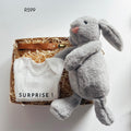A baby gift set in a cardboard box filled with shredded paper, featuring a white baby bodysuit printed with 'SURPRISE!', a brown leather strap bag, and a large gray plush bunny toy. 