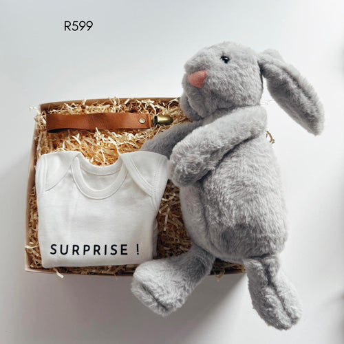 A baby gift set in a cardboard box filled with shredded paper, featuring a white baby bodysuit printed with 'SURPRISE!', a brown leather strap bag, and a large gray plush bunny toy. 
