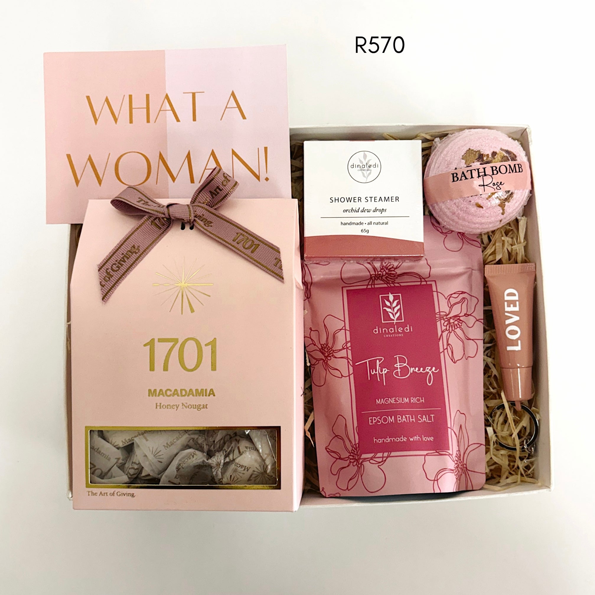 A curated women's self-care gift set in a kraft paper box with shredded filler, featuring a pink macadamia nougat box tied with a brown ribbon, a shower steamer, a bath bomb, and a tube of lotion. A pink card reading 'WHAT A WOMAN!' is placed in the box.