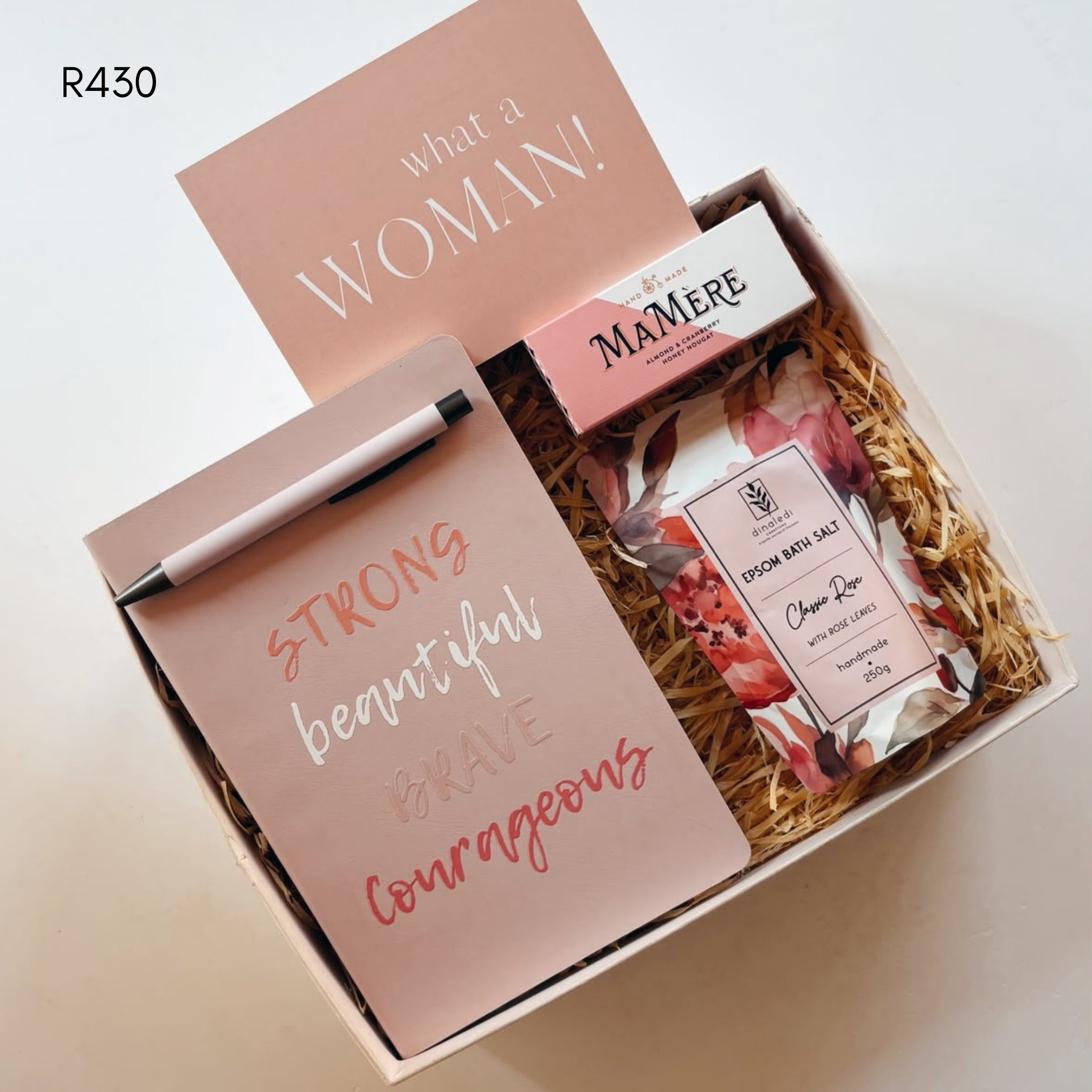 A curated gift box set on a light beige background, featuring a pink card that reads 'what a WOMAN!', a pink notepad with a pen and the words 'STRONG beautiful BRAVE courageous', a box of MAMERE nougat, and a packet of epsom salt nestled in brown paper shred.