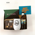 A Father's Day gift set in a black tray with shredded paper filler, featuring a green and black Biltong package, two bags of The Treat Co. mixed nut snacks, a white tumbler printed with 'Dad' in black script, a bottle of Devil's Peak beer, and a blue card that says 'HAPPY FATHER'S DAY'.
