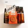 A curated gift basket set containing a Terrodore Gourmet Peanut Butter Cookies box, a bottle of whiskey, a metallic copper tumbler, and branded mixed nuts bags, arranged in a black tray with a congratulatory card.