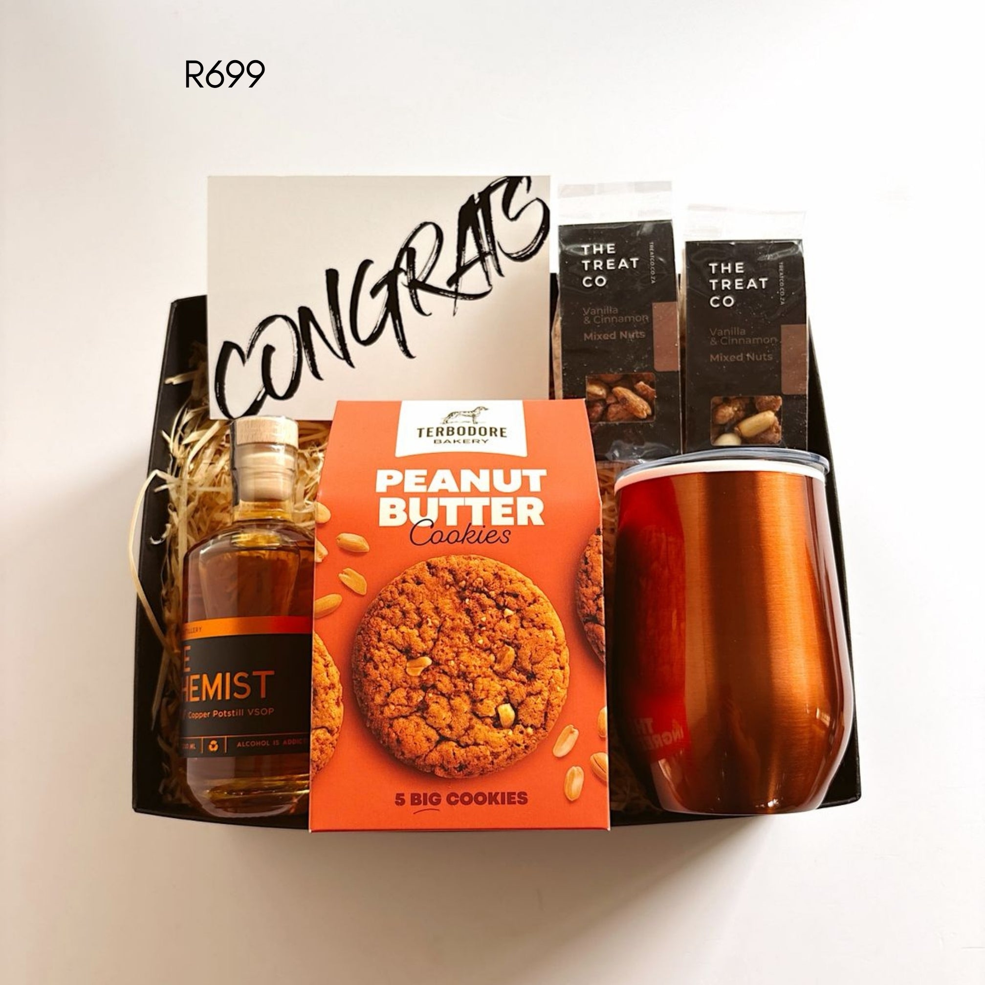 A curated gift basket set containing a Terrodore Gourmet Peanut Butter Cookies box, a bottle of whiskey, a metallic copper tumbler, and branded mixed nuts bags, arranged in a black tray with a congratulatory card.