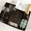 A luxury gift box set containing a bottle of Pierre Jourdan Brut sparkling wine, two ribbed champagne flutes, a box of 1701 Dark Chocolate & Macadamia treats, and a handwritten thank you card, all arranged in a black gift box with black shredded paper.