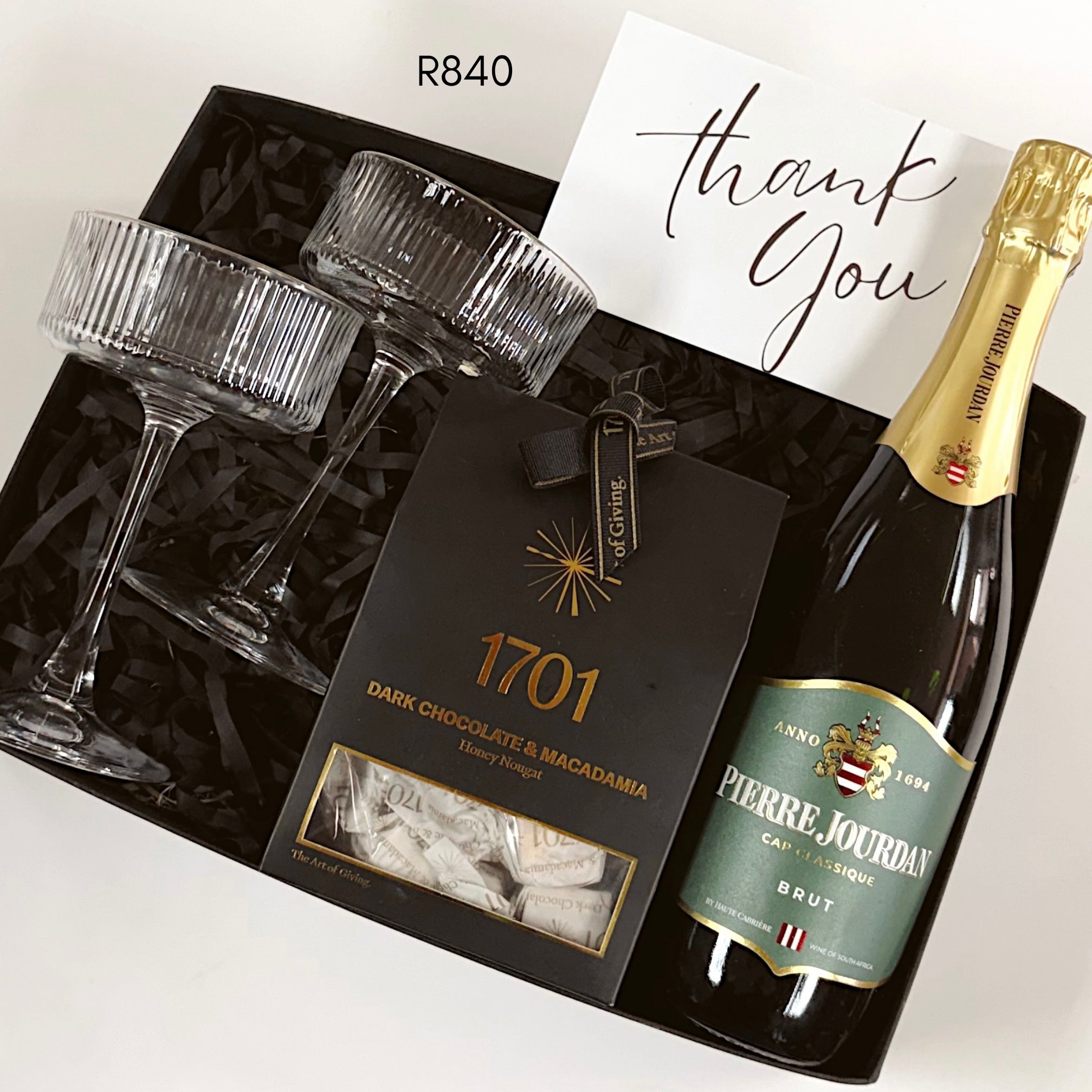 A luxury gift box set containing a bottle of Pierre Jourdan Brut sparkling wine, two ribbed champagne flutes, a box of 1701 Dark Chocolate & Macadamia treats, and a handwritten thank you card, all arranged in a black gift box with black shredded paper.