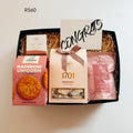 A curated gift box set containing a pink box of Ferrozone Rainbow Unicorn Cookies, a beige box of 1701 Macadamia Honey nougat, a pink bag of Tulip breeze epsom salt, and a small white box of shower steamer, arranged in a black box with shredded paper filler and a 'CONGRATS' card.