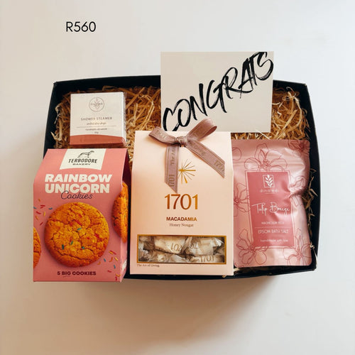 A curated gift box set containing a pink box of Ferrozone Rainbow Unicorn Cookies, a beige box of 1701 Macadamia Honey nougat, a pink bag of Tulip breeze epsom salt, and a small white box of shower steamer, arranged in a black box with shredded paper filler and a 'CONGRATS' card.