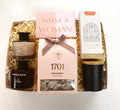 A curated gift box set on a white background, containing a bottle of Bath Salt, a bubble bath product, a pink card reading 'WHAT A WOMAN!', a central 1701 Macadamia nougat, and a Basilei chocolate milo fudge.