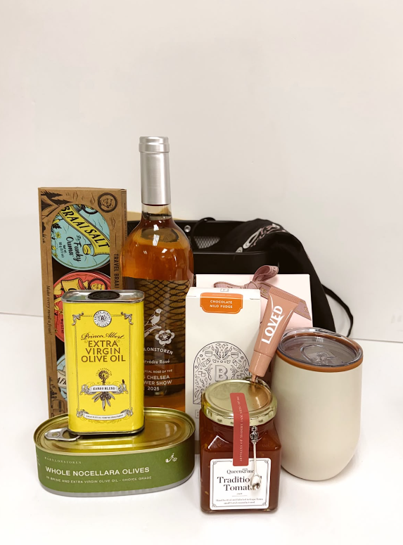 A curated gift basket or food and beverage assortment on a white background, including a bottle of wine, a can of extra virgin olive oil, a tin of whole noccellara olives, a box of spices, a jar of tomato jam, a cream ceramic tumbler, and a black zippered pouch.