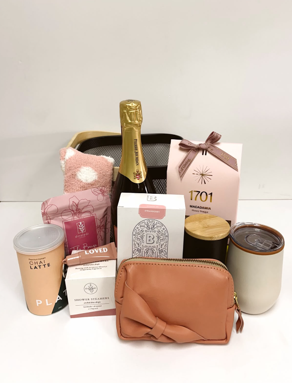A curated gift basket arrangement on a white surface against a white background, featuring a bottle of champagne, a 1701 pink gift box with macadamian nougat, a tan zip pouch with a large bow, a tumbler, packaged snacks, shower steamer and epsom salt.