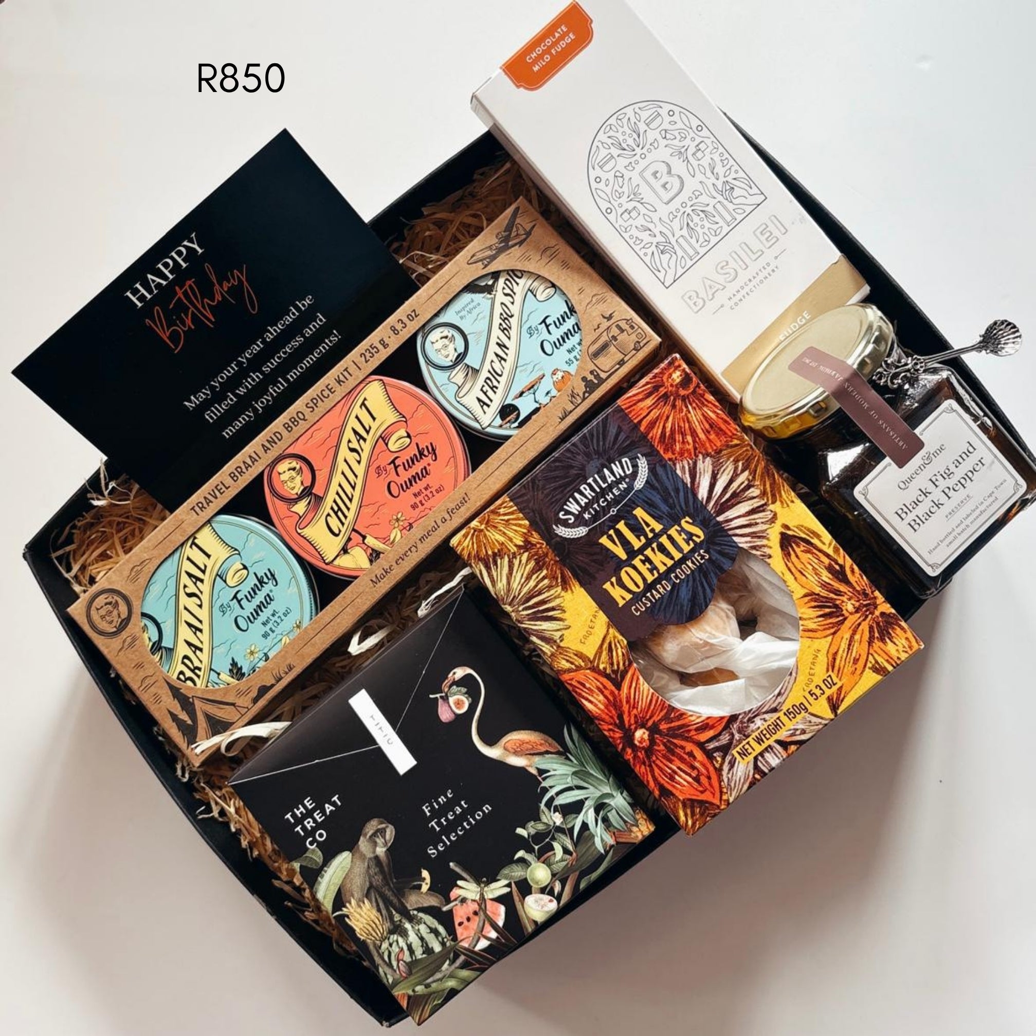 A curated gift box set featuring a black birthday card, chocolate milo fudge, a jar of preserve, and several illustrated three spices, Cla koekies, and fine treats arranged in a black box with shredded paper filler. 