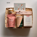 A curated gift box filled with spa and tea items, including a bath salt packet, a tea canister, a box of macadamia nougat labeled 1701, a card reading 'merry merry merry,' and a round bath bomb, arranged in shredded paper on a neutral background.