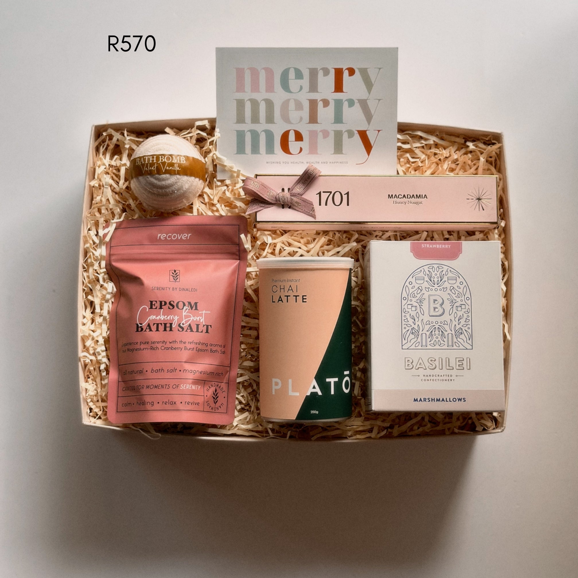 A curated gift box filled with spa and tea items, including a bath salt packet, a tea canister, a box of macadamia nougat labeled 1701, a card reading 'merry merry merry,' and a round bath bomb, arranged in shredded paper on a neutral background.