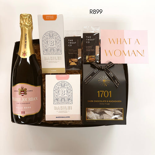 A luxury gift basket containing a bottle of Pierre Jourdan Rosé Champagne, a box of Basilei chocolate milo fudge, mixed nuts treats, a box of Basilei strawberry marshmallows, a bag of 1701 Dark Chocolate & Macadamia nougat, and a pink card that reads 'WHAT A WOMAN!'.