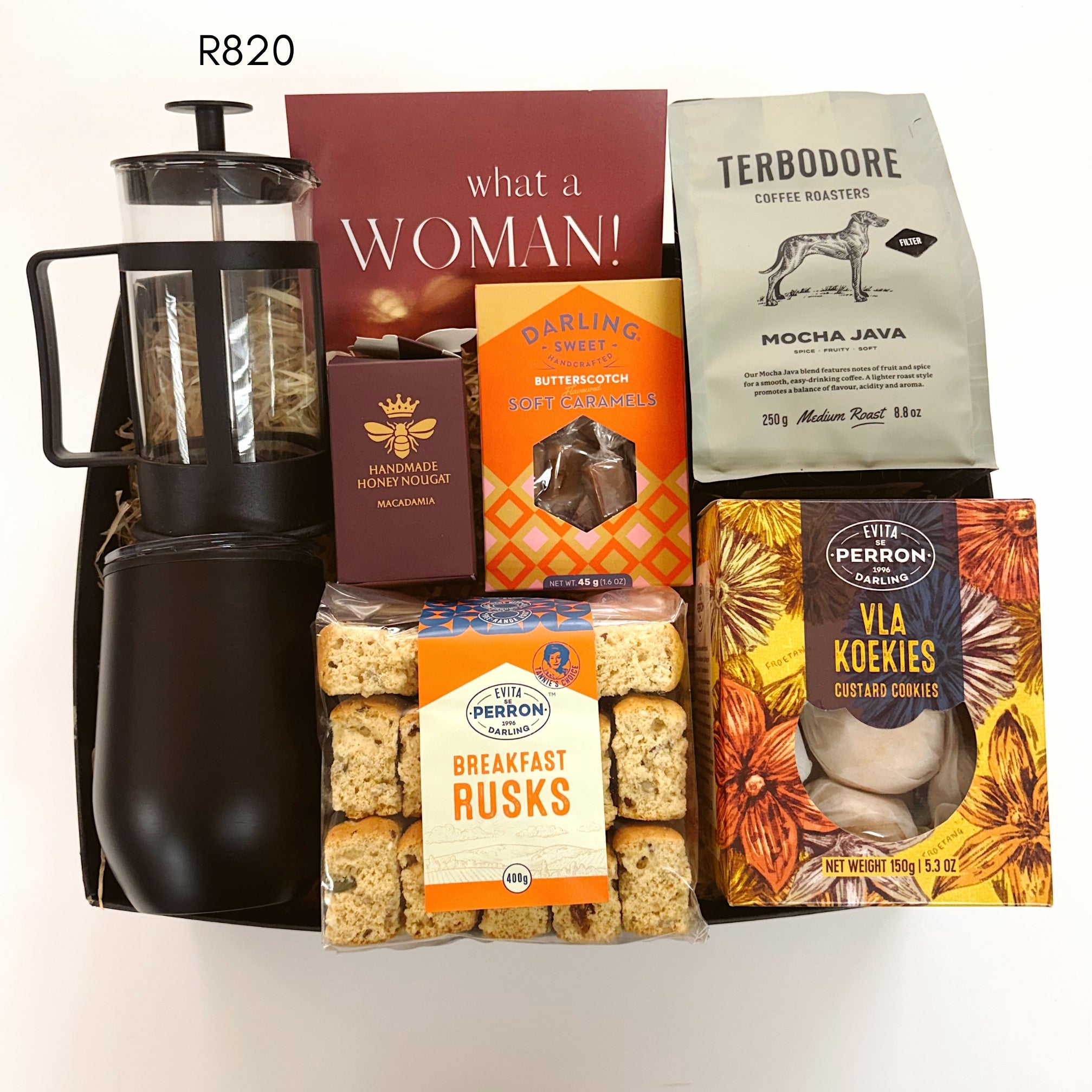 A curated gift basket or hamper containing multiple packaged food and beverage items arranged in a black tray against a plain white background. Visible items include a black French press coffee maker, a red card titled 'what a WOMAN!', a bag of Terbodore Coffee Roasters Mocha Java, a box of honeycomb, a bag of breakfast rusks, and a box of Vla Koekies cookies.