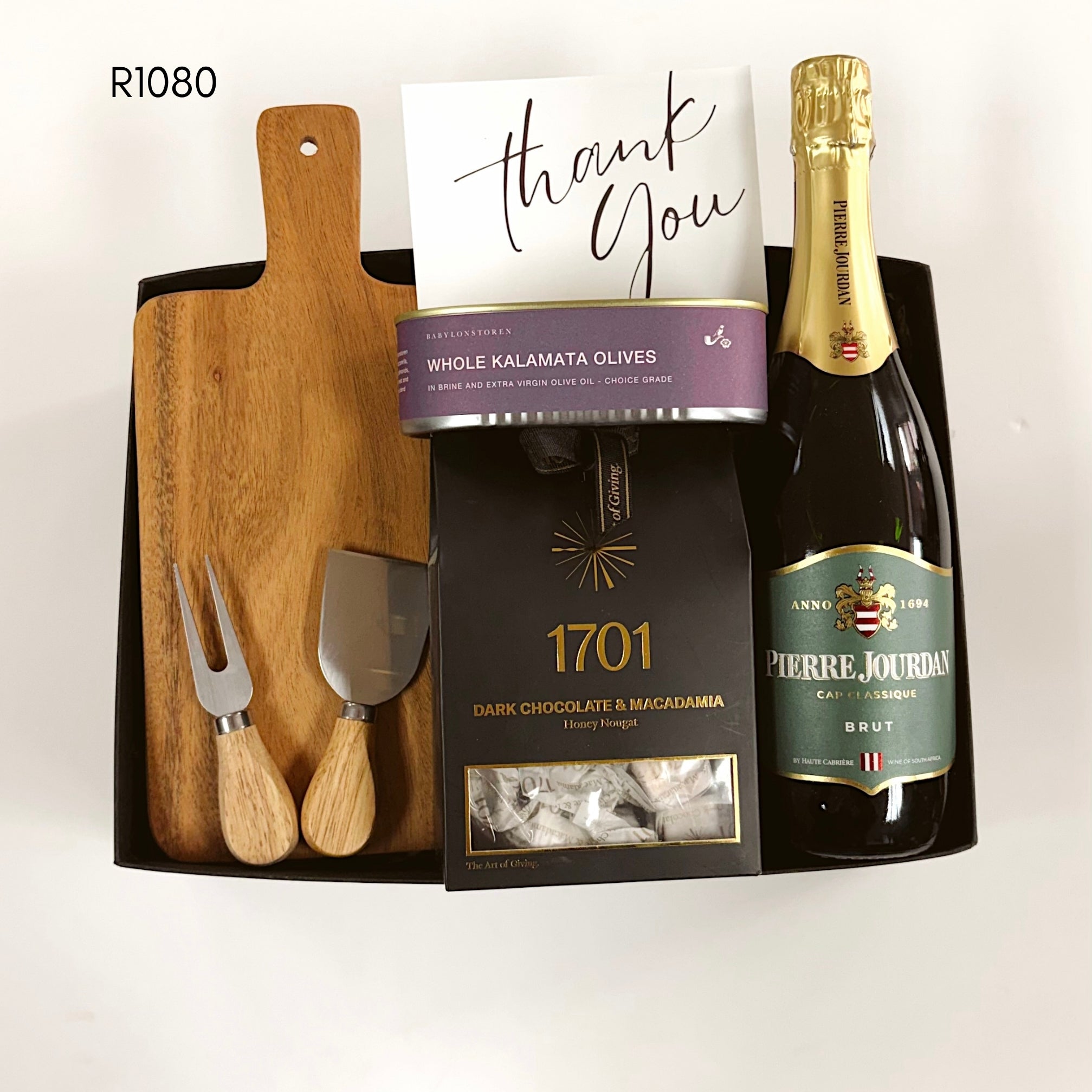 A curated gift basket set containing a wooden cutting board, two wooden-handled serving cheese knifes, a bottle of Pierre Jourdan Brut sparkling wine, a box of 1701 dark chocolate and macadamia nougat, and a tin of whole kalamata olives, arranged in a black tray with a handwritten thank you card.