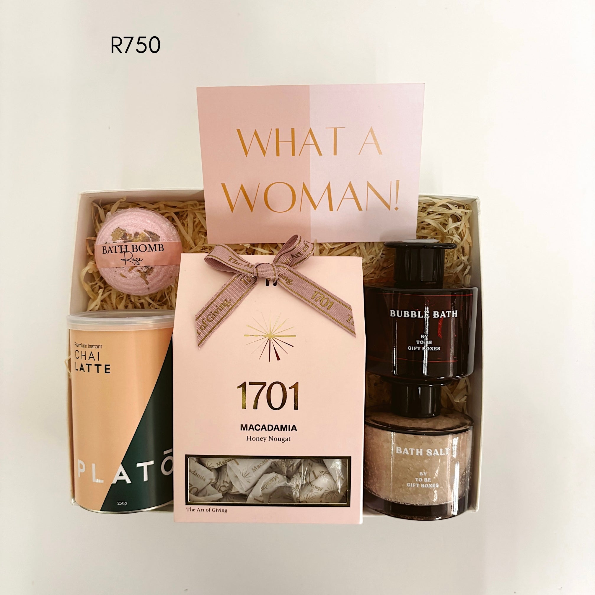 A curated pamper gift set in a box labeled 'WHAT A WOMAN!' with a pink card, a bath bomb, a Plato tin of chai latte, Macadamia nougat, and bubble bath items, photographed on a plain light background.
