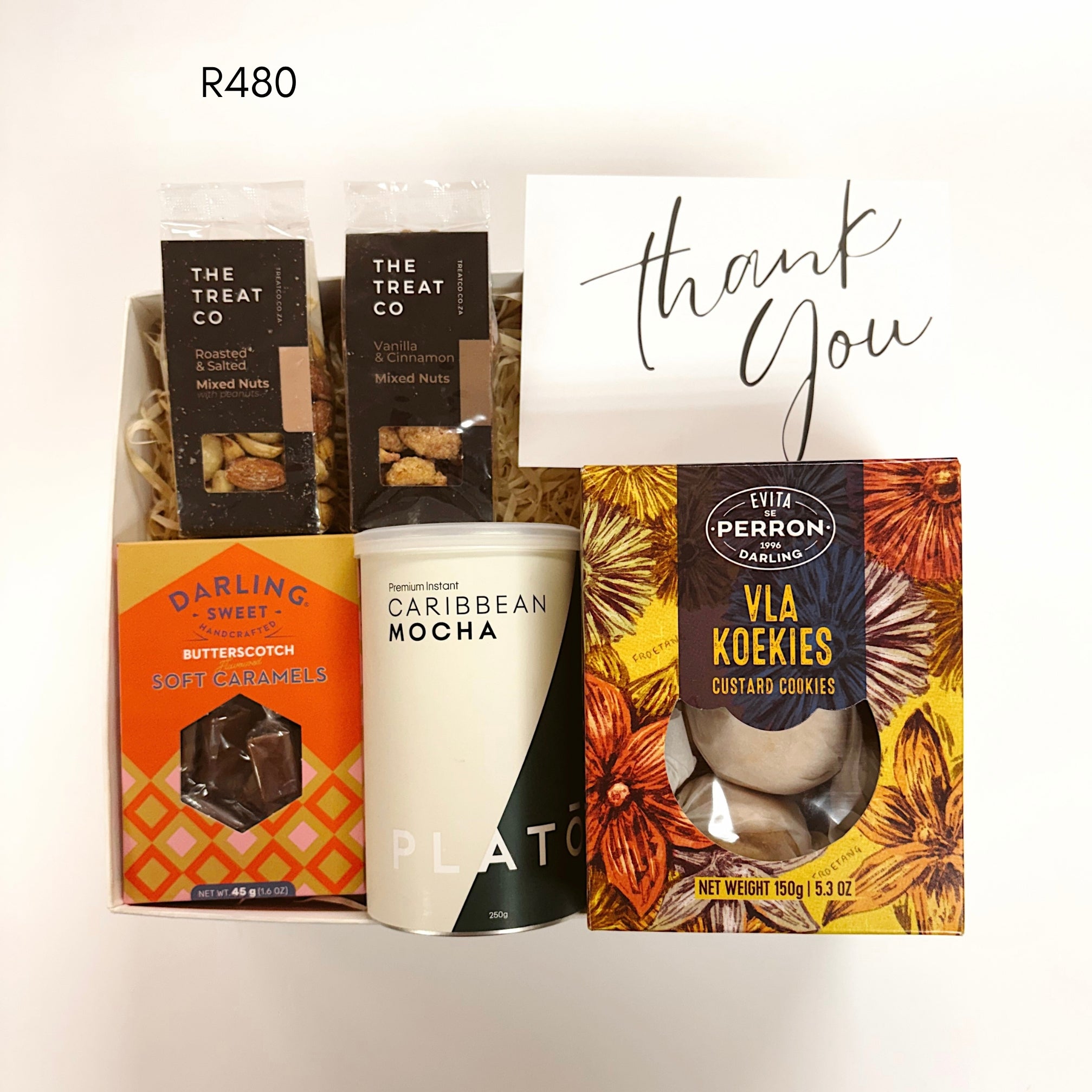 A curated gift basket set featuring a can of Plato Caribbean Mocha, a box of Vla Koekies, a box of Darling Sweet Butterscotch soft caramels, and two bags of The Treat Co mixed nuts, arranged with a white 'thank you' card on a white background.