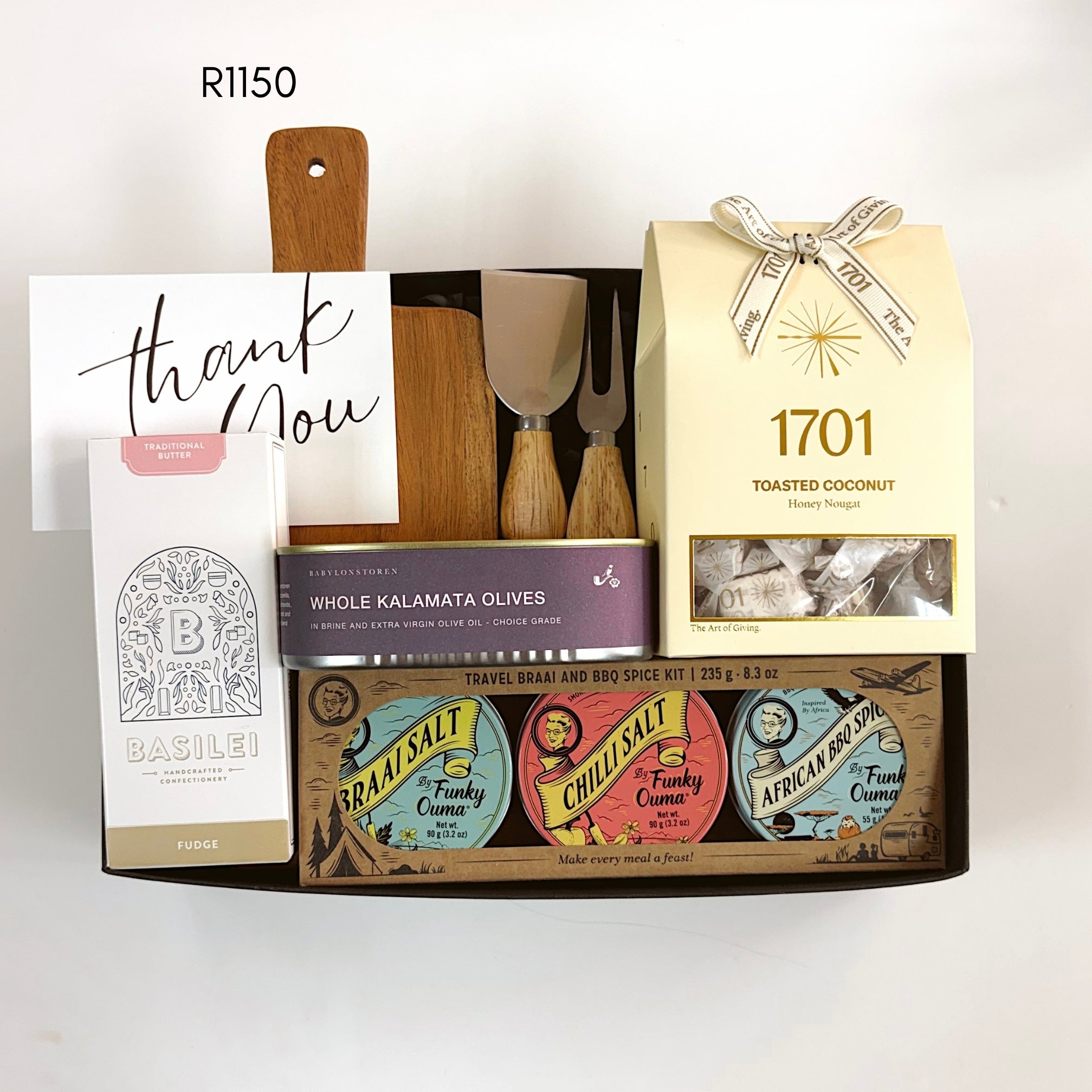 A curated gift basket set containing a boxed set of Basilei traditional butter, a box of 1701 toasted coconut nougat, a purple tin of whole kalamata olives, and a collection of three colorful spices. The items are arranged in a wooden tray with wooden cheese utensils and a small wooden cutting board, with a 'thank you' card on the left.