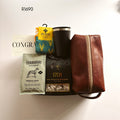 A curated gift set arranged on a white background, featuring a brown leather zip pouch, a black insulated tumbler, a bag of Terbodore coffee, a box of 1701 dark chocolate and magadama nougat, and a teal and yellow set of socks.