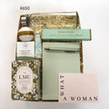A curated gift box set filled with shredded paper, featuring Jasmine and orange blossom tea, a glass bottle, a bath bomb, a shower steamer, a notebook and pen, salted caramel, and a pink card that reads 'WHAT A WOMAN'.