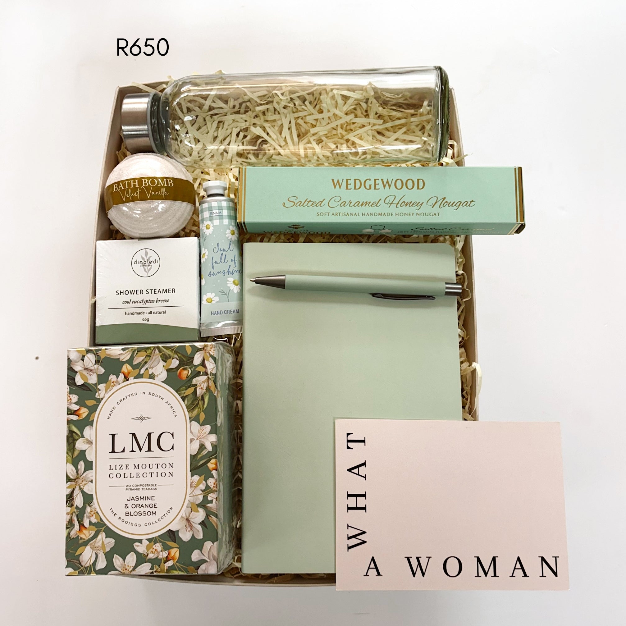 A curated gift box set filled with shredded paper, featuring Jasmine and orange blossom tea, a glass bottle, a bath bomb, a shower steamer, a notebook and pen, salted caramel, and a pink card that reads 'WHAT A WOMAN'.