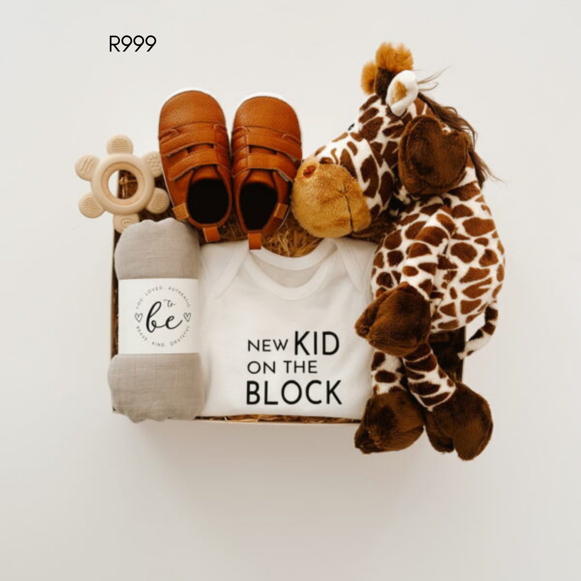 A baby gift set arranged in a box on a white background, including a white onesie printed with 'NEW KID ON THE BLOCK', a pair of brown baby shoes, a gray blanket with a 'to be' label, a wooden teething ring, and a plush giraffe toy. 