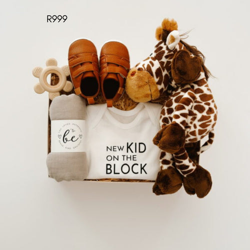 A baby gift set arranged in a box on a white background, including a white onesie printed with 'NEW KID ON THE BLOCK', a pair of brown baby shoes, a gray blanket with a 'to be' label, a wooden teething ring, and a plush giraffe toy. 