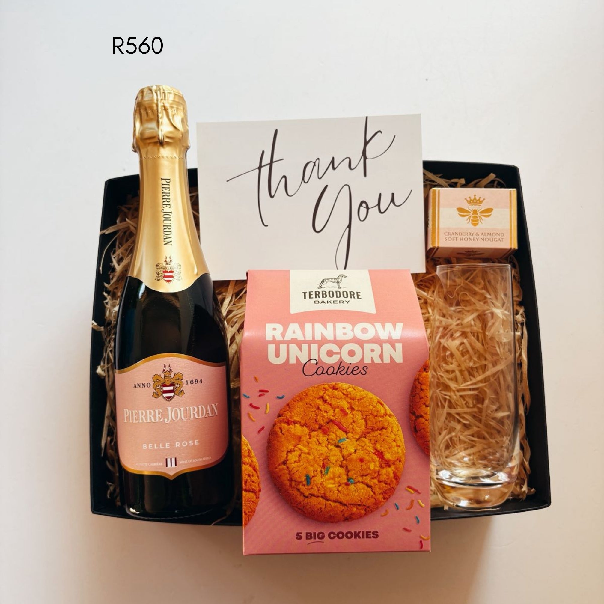A curated gift basket set containing a bottle of Pierre-Jourdan Sparkling Wine, a box of TERBODORE Rainbow Unicorn Cookies, nougat, a clear glass, and a handwritten thank you card, arranged in a black tray with shredded paper filler.